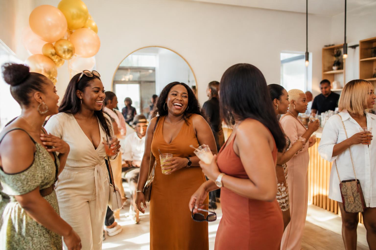 Friends laughing together at a relaxed birthday gathering with gold and blush balloons in a bright cream and oak event space